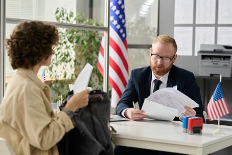 A woman and a man sit at a desk with two US flags prominently displayed.